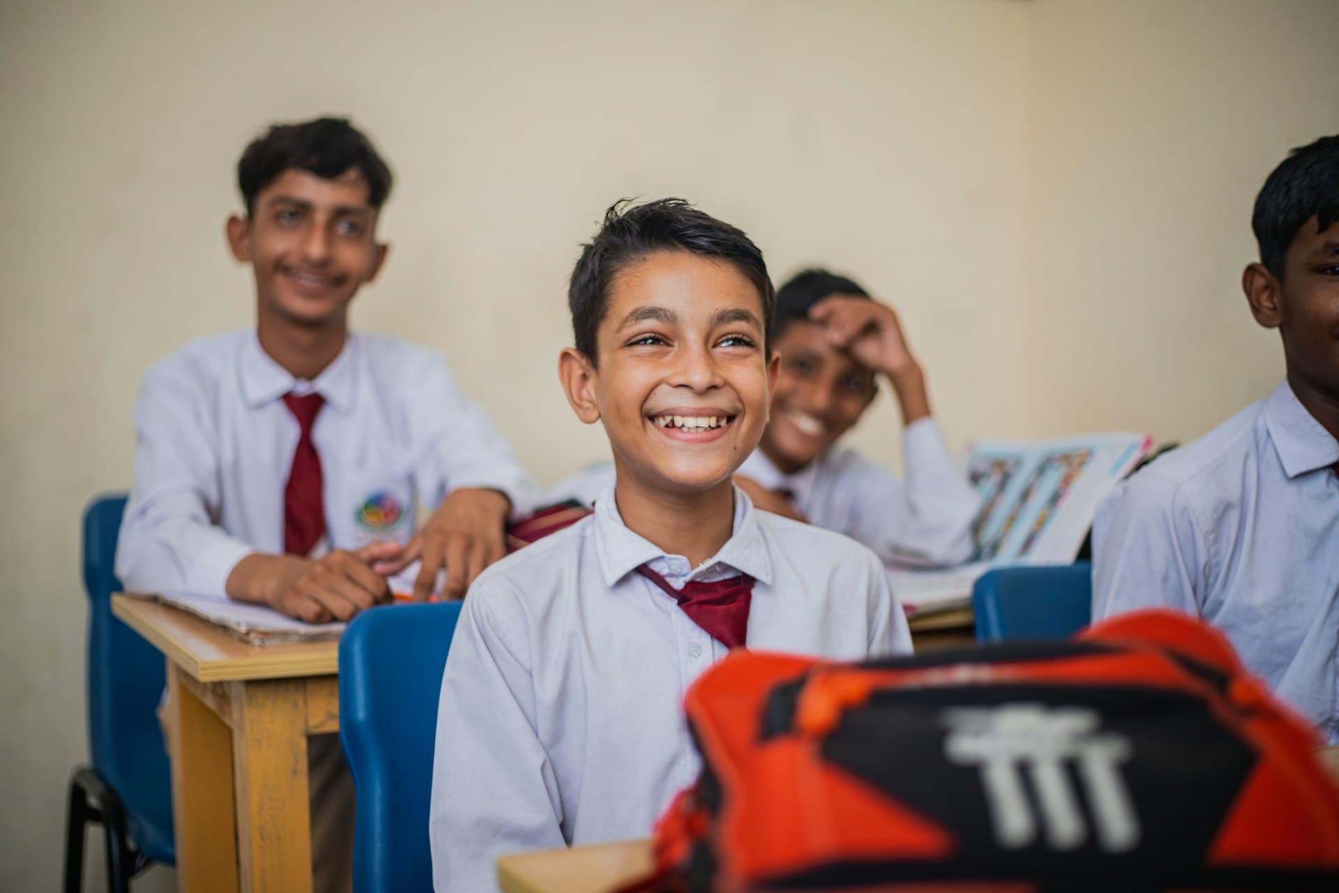 A smiling student at his desk in class, with classmates laughing behind him